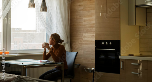 Woman thoughtfully looking at window with cup after reading mail at table in kitchen
