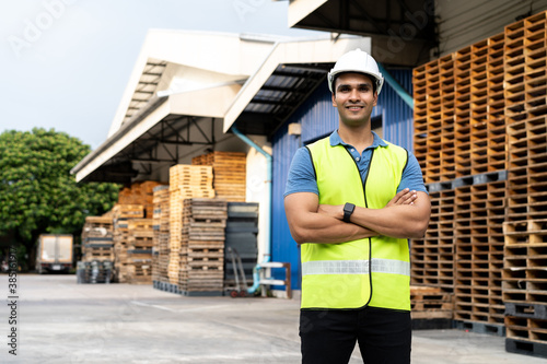 Portrait of young Indian worker with arm folded working in logistic industry outdoor in front of factory warehouse. Smiling happy man in hard hat looking at camera arms crossed at depot