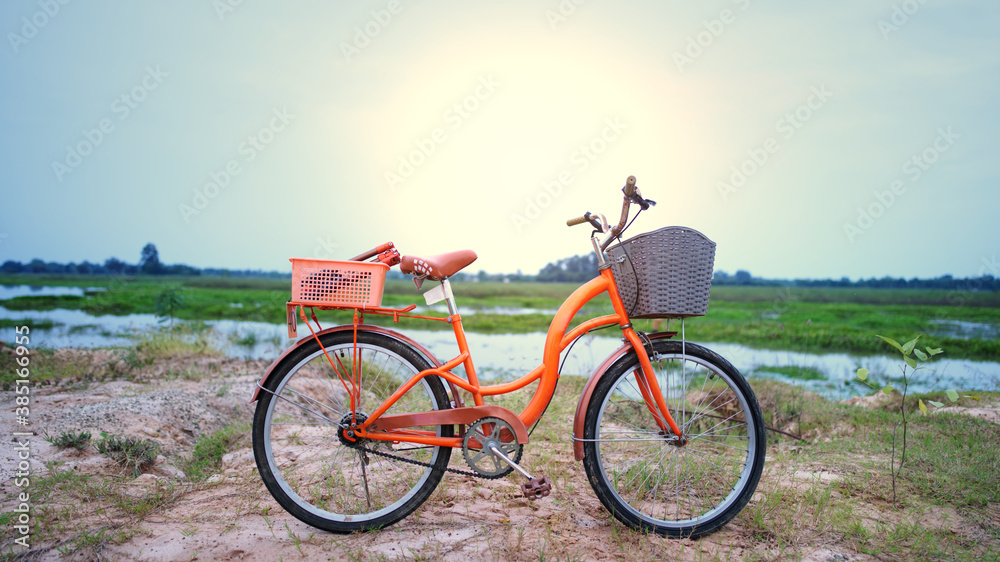 An orange retro-style bicycle with a basket for luggage, parked by the river as the sun shines through the concept of exercise and leisure with leisure activities on the weekend.
