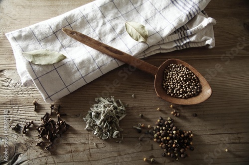 Coriander (Coriandrum sativum), Peppercorns (Piper nigrum), Sage (Salvia) and dried Cloves (Syzygium aromaticum) with a wooden spoon and a kitchen towel on a rustic wooden surface photo