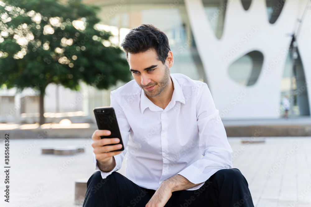 Handsome male entrepreneur using smart phone sitting against built structure in city Handsome male entrepreneur using smart phone sitting against built structure in city