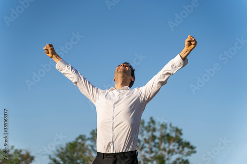 Successful businessman with arms raised standing against clear blue sky