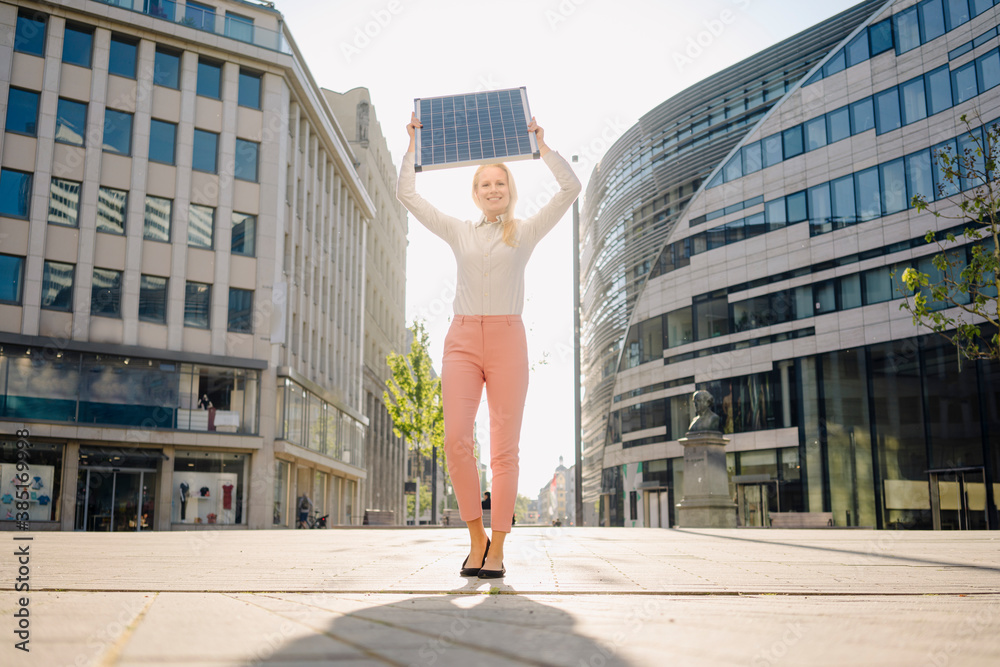 Smiling beautiful female professional holding solar panel while standing on footpath in financial district Smiling beautiful female professional holding solar panel while standing on footpath in financial district