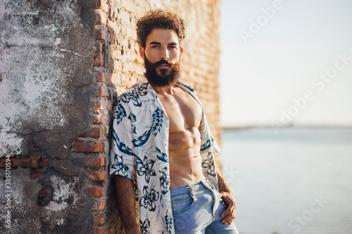 Young bearded man standing with shirt open against brick wall photo