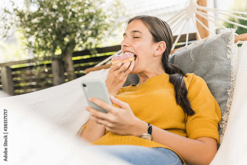 Young woman eating donut while using smart phone on hammock in yard Young woman eating donut while using smart phone on hammock in yard