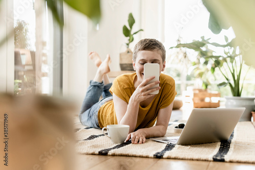 Woman using mobile phone while lying by coffee cup and laptop on carpet at home