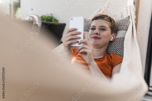 Mid adult woman with wireless headphones using smart phone while lying on hammock in porch photo