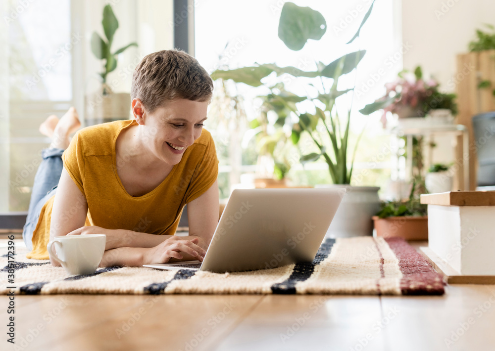 © UWE_UMSTAETTER/Westend61 - Smiling mid adult woman with short hair using laptop while relaxing on carpet at home © UWE_UMSTAETTER/Westend61 - Smiling mid adult woman with short hair using laptop while relaxing on carpet at home