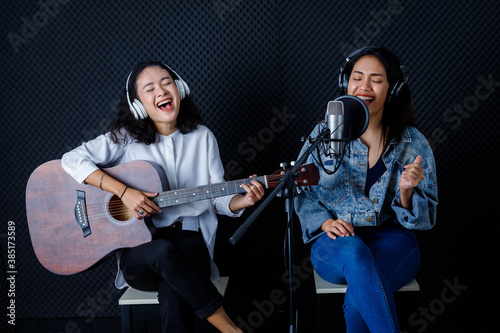 Happy cheerful pretty smiling of portrait of Two young Asian woman vocalist Wearing Headphones with a guitar recording a song front of microphone in a professional studio