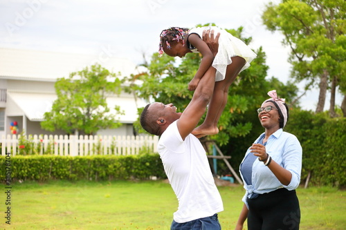 African American family playing with young happy little daughter on green grass field while enjoying summer garden outside the house in the neighborhood with copy space