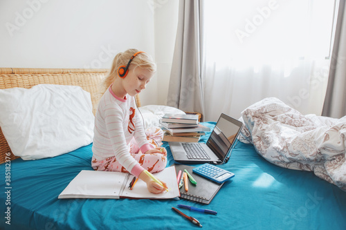 Caucasian girl child sitting in bed and learning online on laptop Internet. Virtual class lesson on video during self-isolation at home. Distant remote video education. Homeschooling nowdays.