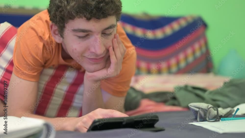 Home scenes: a nice Caucasian boy studies on his parents' bed, in reality he is completely absorbed by his smartphone. In front of him, the notebook left open is not the object of his attention. Home scenes: a nice Caucasian boy studies on his parents' bed, in reality he is completely absorbed by his smartphone. In front of him, the notebook left open is not the object of his attention.