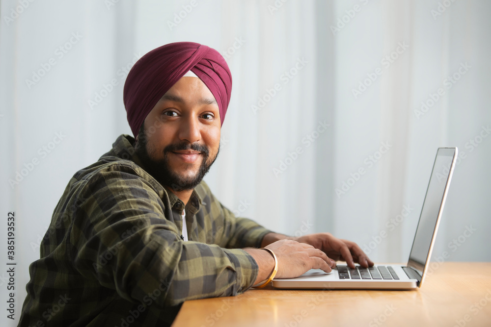 A SIKH MAN LOOKING AT CAMERA AND SMILING WHILE AT WORK WITH LAPTOP A SIKH MAN LOOKING AT CAMERA AND SMILING WHILE AT WORK WITH LAPTOP