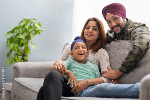 A SIKH BOY SMILING AND LEANING OVER MOTHER SITTING ON SOFA WHILE FATHER BENDS OVER TO THEM AND SMILES	 photo