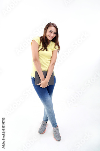 Yong Asian youth girl in casual dress point holding book notebook look at camera stand pose cross legged on white background