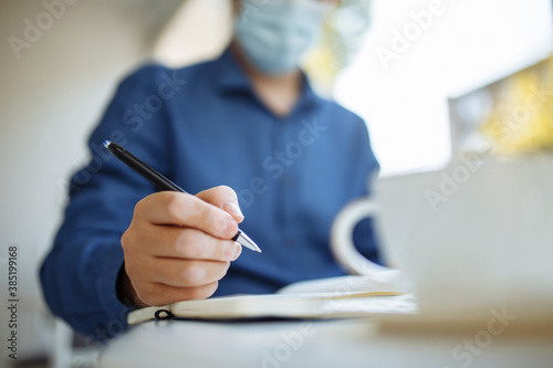Young businessman wearing a medical mask sits in a cafe with a laptop working and taking notes. Coronavirus pandemic, distant work and business concept.