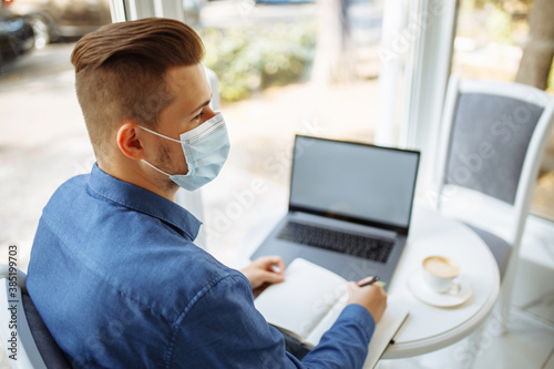 Young businessman wearing a medical sterile mask taking noted in a cafe in front of a laptop waiting for a cup of coffee. Coronavirus pandemic qurantine and wokring remotely concept. photo