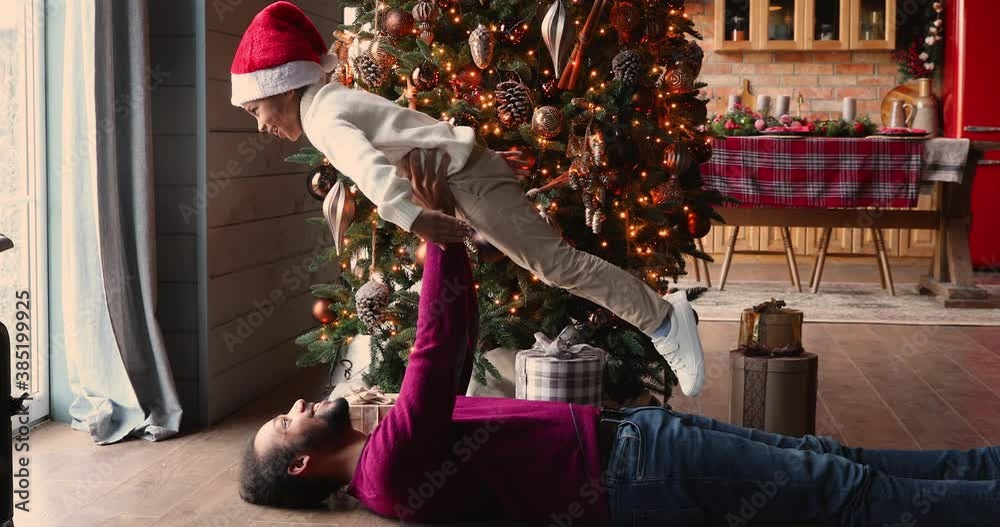 African ethnicity father lying on warm wooden floor in decorated with lights and Christmas tree living room holding lifting up preschool son, family having fun playing at home, happy holidays concept