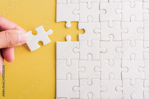 Top view flat lay of hand-holding last piece white paper jigsaw puzzle game last pieces put to place for solve problem complete mission, studio shot on a yellow background, quiz calculation concept