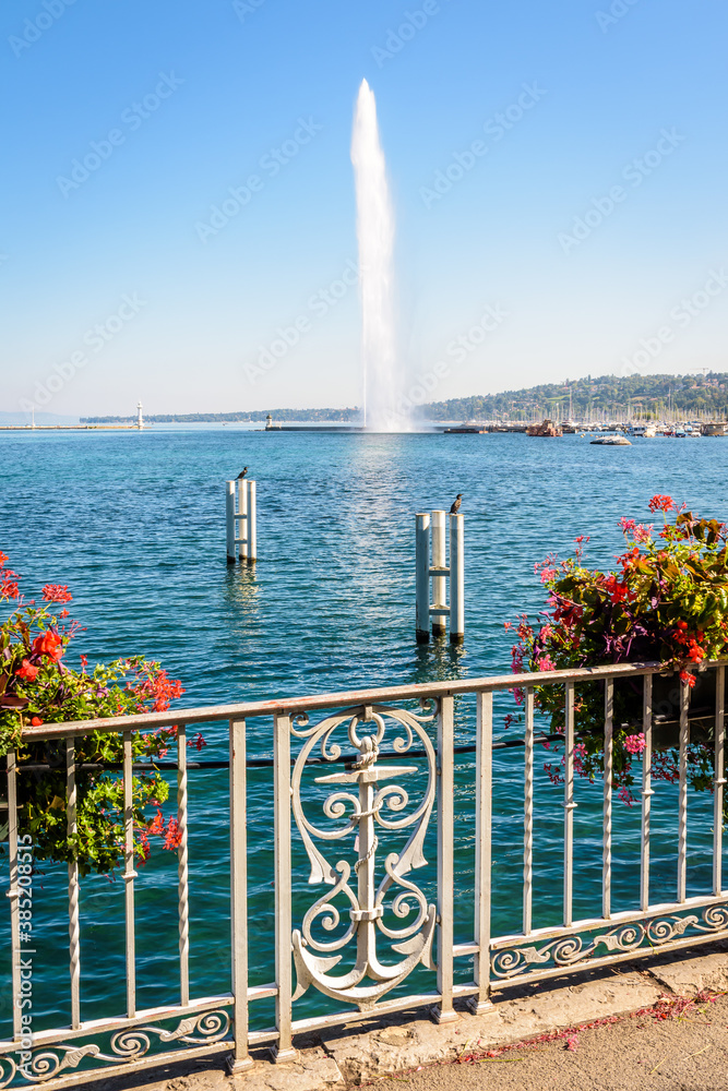 Φωτογραφία The Jet d'Eau, the 140 meter-high water jet fountain in the bay of Geneva, Switzerland, seen from the Jardin Anglais with a wrought iron railing with anchor and potted geraniums by a sunny summer day