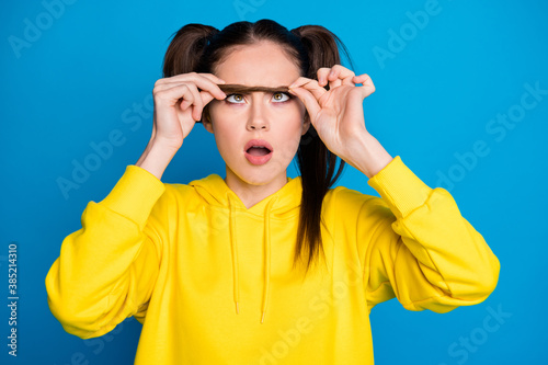 Photo of pretty shocked stupor lady hold tail making false one eyebrow open mouth need correction grooming wear casual yellow hoodie pullover isolated bright blue color background