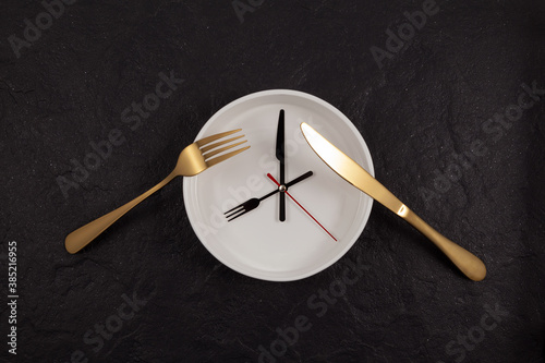 White deep plate, knife and fork. Clock on a dark textured stone background with copy space. The hands point to 8 o'clock. Concept - interval fasting or autophagy photo