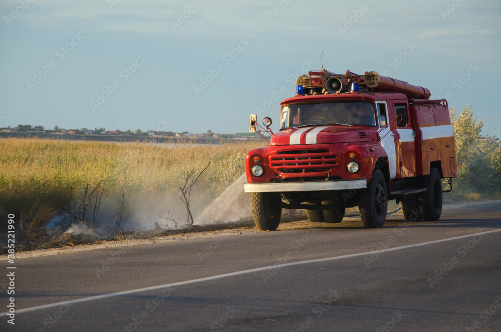 An old firetruck extinguishes a fire near the road. The grass is burning near the village.