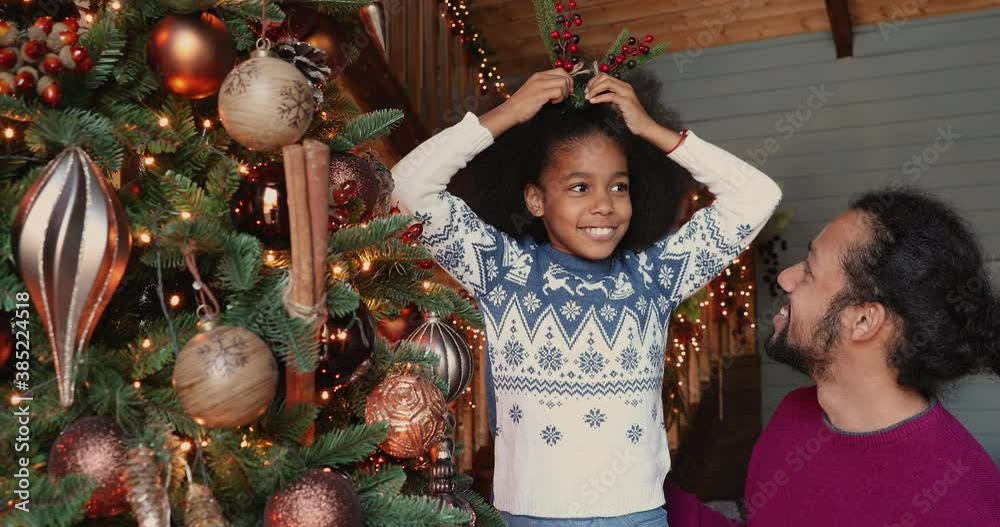 African kid girl play during Christmas tree decoration, hangs spruce twigs on artificial fir, talk to father enjoy New Year preparation create festive atmosphere at home. Happy winter holidays concept