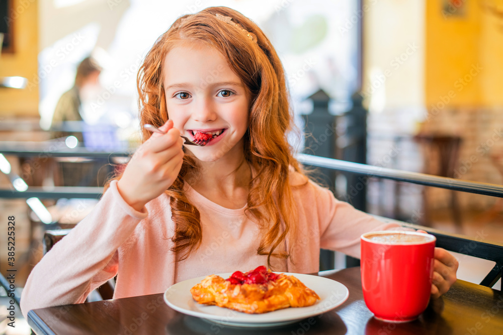 Little red-haired girl eating cake in cafe, smiling looking into the distance. Little red-haired girl eating cake in cafe, smiling looking into the distance.