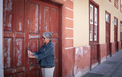 One senior caucasian woman with gray hair tries to open an old red door closed by lock. Traditional spanish home with red windows and doors made by wood. Rustic style. © luciano