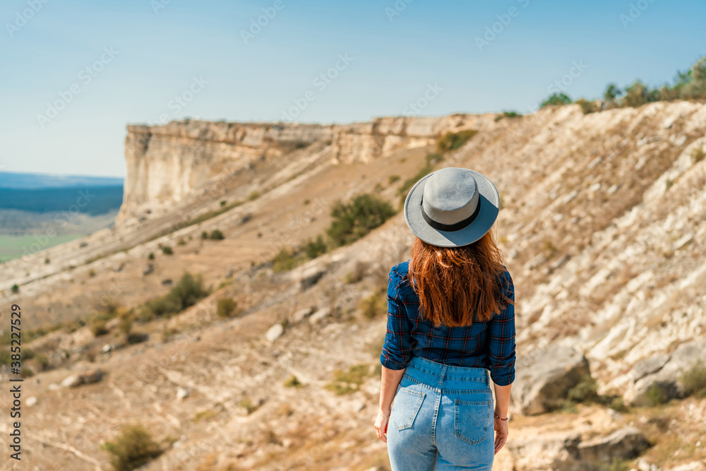 Naklejka premium Rear view of a young woman in a plaid shirt and hat standing on a mountain and admiring the landscape, White rock in the Crimea