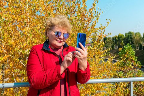 Caucasian senior woman in red jacket using smartphone, read e-book, leasten music or take online education in park in autumn weather Video call, chat Distance meeting  photo