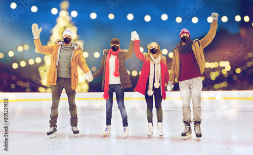 friendship, gesture, christmas and leisure concept - friends wearing face protective masks for protection from virus disease waving hands at outdoor skating rink over holiday lights background