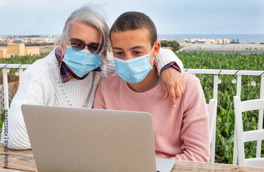 Senior grandmother and teenage grandson looking at laptop together outdoors on the terrace. They both wear a surgical mask to avoid coronavirus infection Senior grandmother and teenage grandson looking at laptop together outdoors on the terrace. They both wear a surgical mask to avoid coronavirus infection