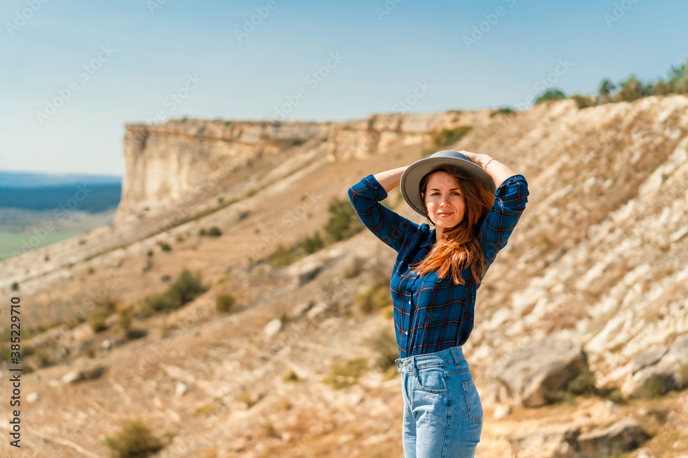 Naklejka premium A young woman in a plaid shirt and hat stands on a mountain with a view of the rustic landscape, White rock in the Crimea