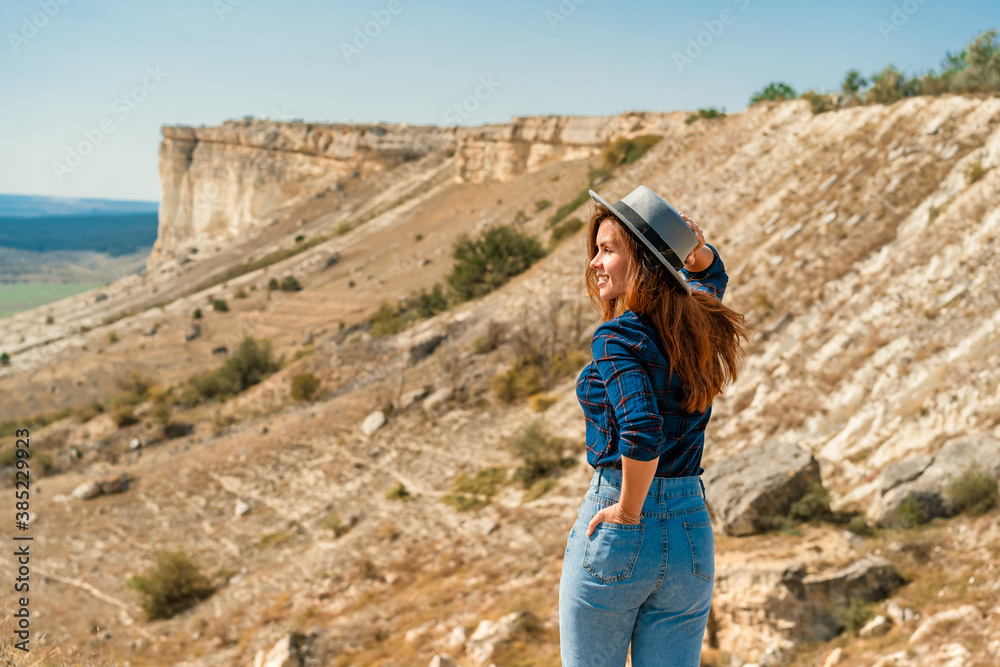 A young woman in a plaid shirt and hat stands on a mountain with a view of the rustic landscape, White rock in the Crimea