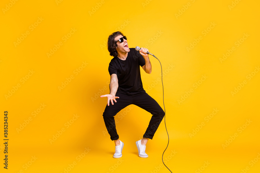 Full length photo of young man dance hold mic sing open mouth wear black t-shirt pants white sneakers sunglass isolated yellow color background