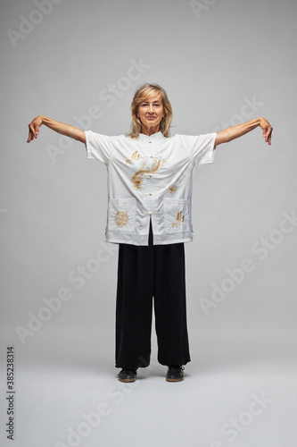 Mature blonde woman practicing Chi kung and Tai Chi on a white background. She wears a traditional white chinese Tai Chi jacket, black trousers and black shoes with ying yang symbol