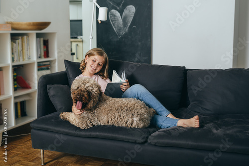 teenager girl sitting on sofa with her dog at home and reading book