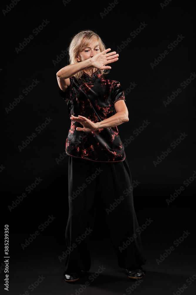 Obraz premium Mature blonde woman practicing Tai Chin Chen style in a black background wearing a traticional chinese black jacket with red decoration, black trousers and traditional shoes with ying yang symbol