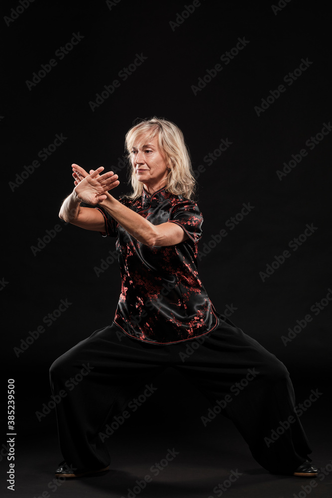 Mature blonde woman practicing Tai Chin Chen style in a black background wearing a traticional chinese black jacket with red decoration, black trousers and traditional shoes with ying yang symbol