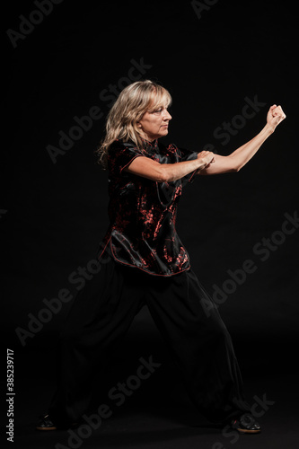 Mature blonde woman practicing Tai Chin Chen style in a black background wearing a traticional chinese black jacket with red decoration, black trousers and traditional shoes with ying yang symbol