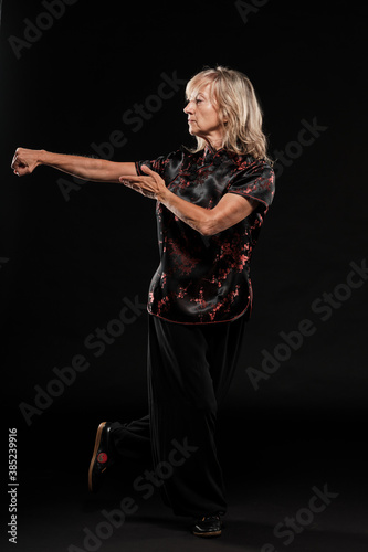 Mature blonde woman practicing Tai Chin Chen style in a black background wearing a traticional chinese black jacket with red decoration  black trousers and traditional shoes with ying yang symbol