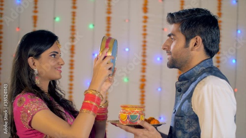 Newly-wed Indian couple celebrating Karwa Chauth festival in traditional wear. Beautiful wife happily looking at the moon and then to her handsome husband through a sieve - Karwa Chauth rituals photo