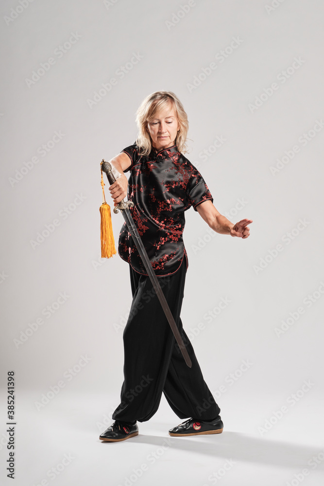 Fototapeta premium Mature blonde woman practicing Tai Chin with sword in a white background wearing a traditional chinese black jacket with red chinese decoration, black trousers and chinese shoes with ying yang symbol