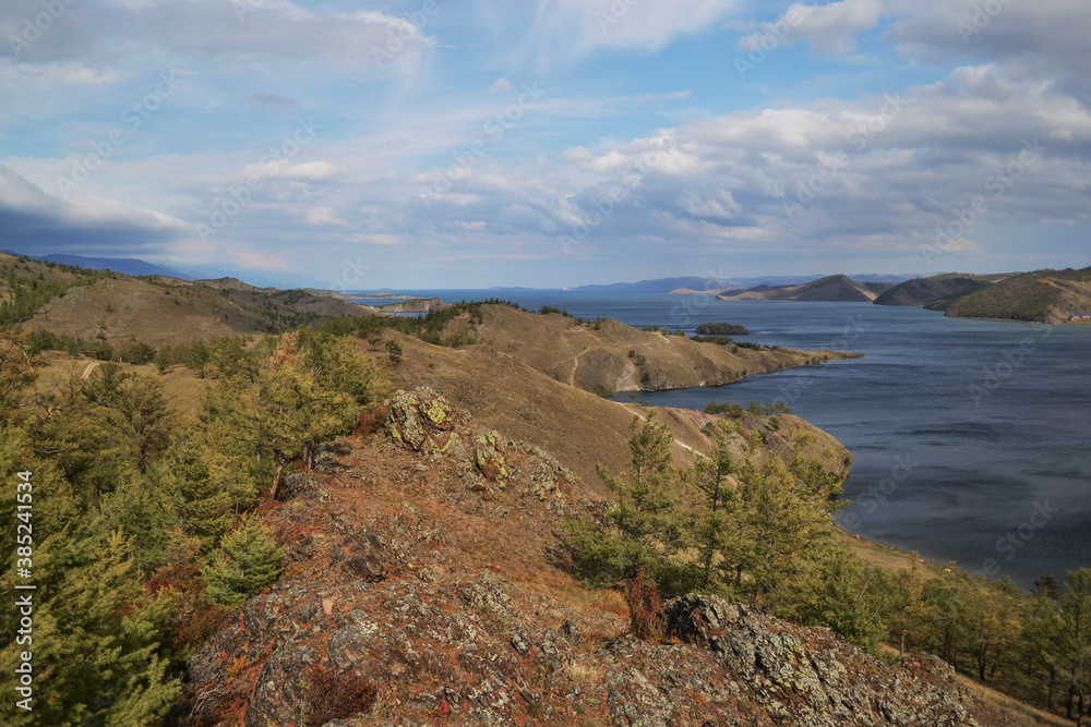 Autumn view of landscape of Lake Baikal with islands and peninsulas and mountains on horizon. Blue water, green trees, clouds in sky Autumn view of landscape of Lake Baikal with islands and peninsulas and mountains on horizon. Blue water, green trees, clouds in sky