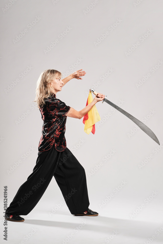 Mature blonde woman practicing Tai Chin with saber in a white background wearing a traditional chinese black jacket with red chinese decoration, black trousers and chinese shoes with ying yang symbol Mature blonde woman practicing Tai Chin with saber in a white background wearing a traditional chinese black jacket with red chinese decoration, black trousers and chinese shoes with ying yang symbol