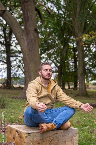 Photo of a young and attractive man sitting on a tree trunk practising meditation in the middle of nature. Self isolation in a forest. Relax
