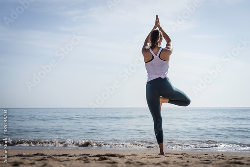 Sporty young woman doing Yoga exercises using a gym mat along the beach in Lisbon, Portugal. Playful woman working as freelance Yoga teacher doing fitness workout on the beach at sunset. Healthy life