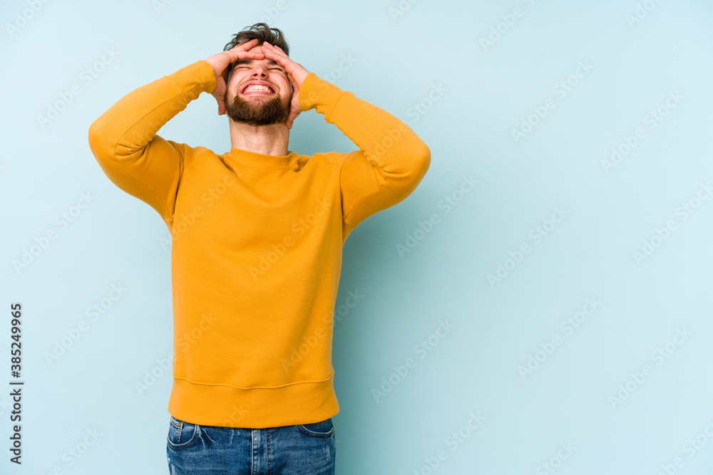 Naklejka premium Young caucasian man isolated on blue background laughs joyfully keeping hands on head. Happiness concept.